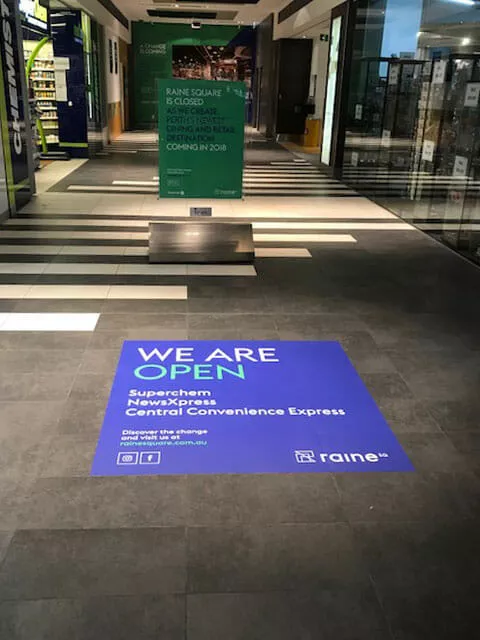 Shopping mall hallway with a blue floor sign reading 'We Are Open: Superchem, NewsXpress, Central Convenience Express.' Bright, welcoming atmosphere