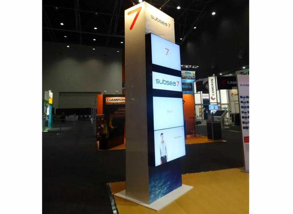 Tall display structure at a trade show booth featuring the brand "Subsea 7" with several illuminated screens, against a backdrop of various exhibition stands