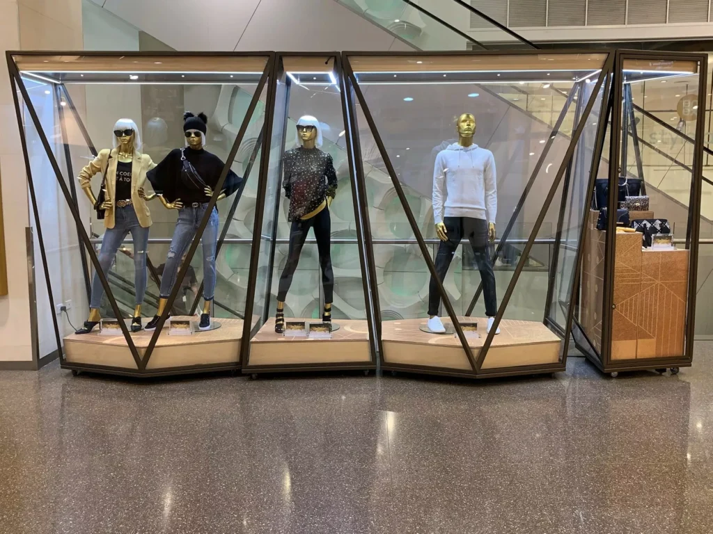 Four mannequins dressed in stylish, casual outfits, displayed in a geometric glass case inside a modern shopping mall