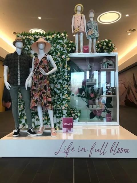Mannequins in floral outfits and hats stand in front of a lush floral backdrop, promoting "Life in Full Bloom" at Kmart, evoking a spring theme