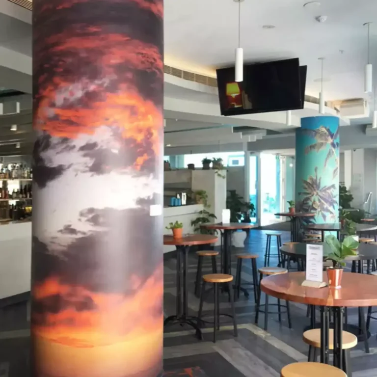 Modern cafe interior with scenic column art depicting a vivid sunset and palm trees. Wooden tables, stools, and potted plants create a cozy ambiance.