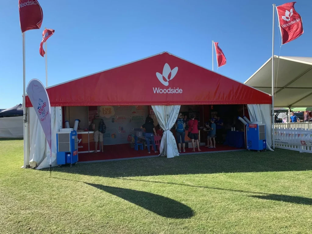 Red Woodside tent at an outdoor event on a sunny day. People gather inside and near the entrance. Flags with the Woodside logo stand tall.