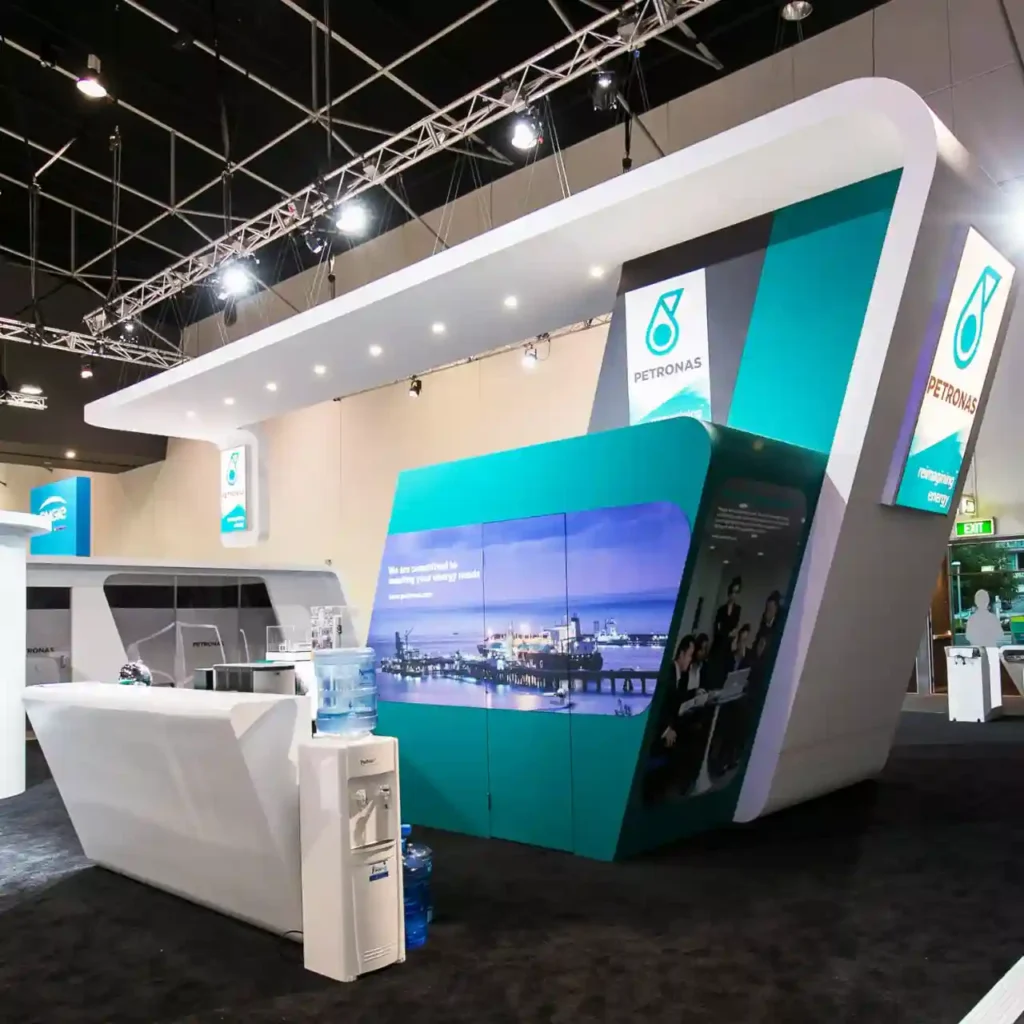 Modern exhibition booth for Petronas, featuring blue and green branding, digital screens displaying an industrial scene, and a sleek reception counter.
