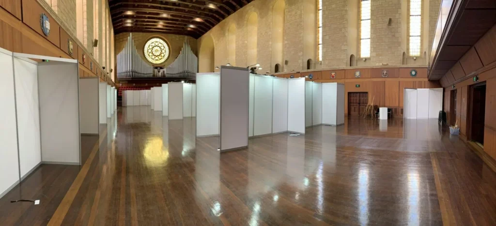 Spacious hall with polished wooden floors and high ceiling. White exhibition booths line the sides, and a large ornate window is at the back, creating a serene atmosphere