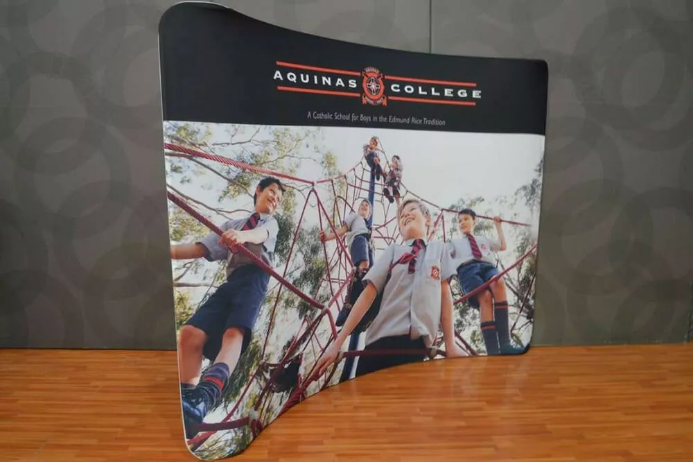 Children in school uniforms are joyfully climbing a red jungle gym against a backdrop of trees. A banner above reads “Aquinas College.”