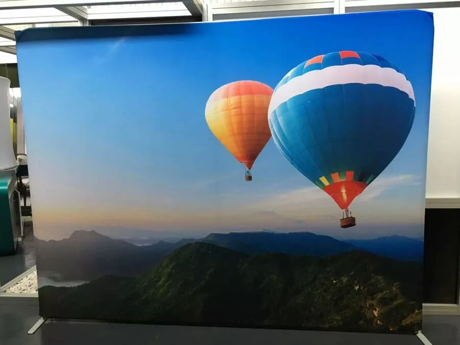 Two colorful hot air balloons, one orange and one blue, float over a lush green mountain range under a clear blue sky portable display