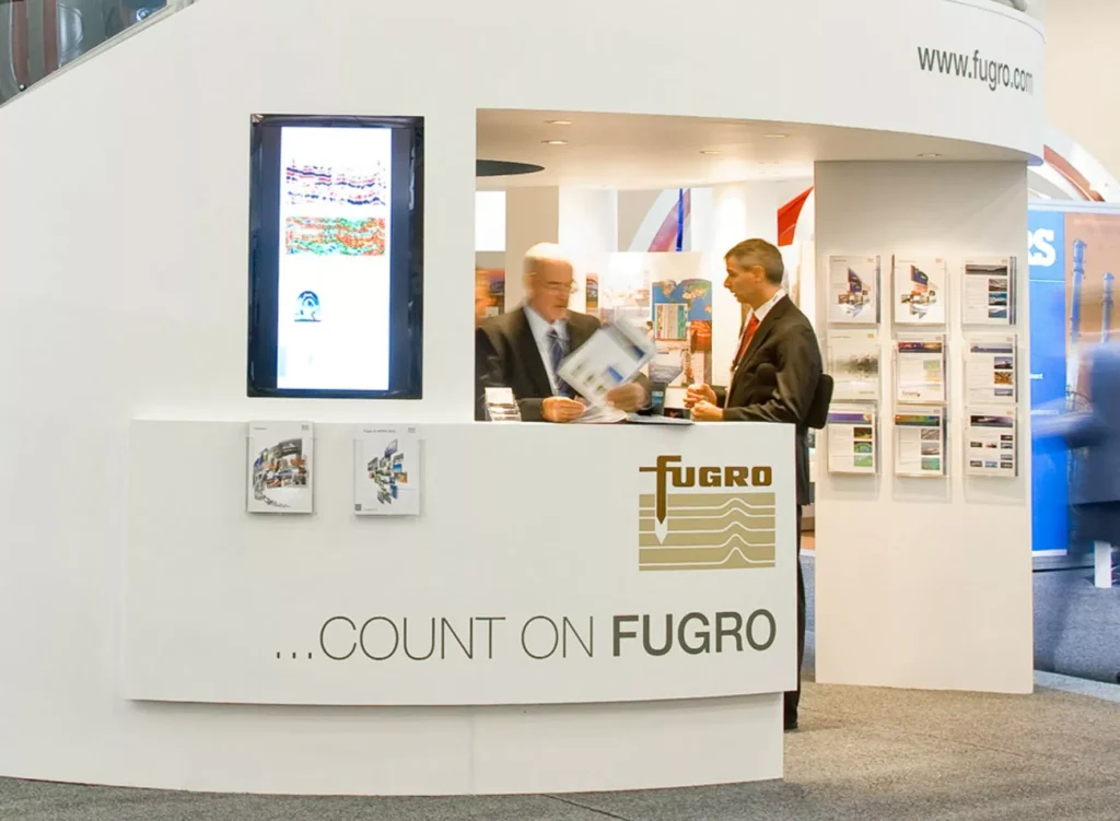 Booth with "FUGRO" branding at an exhibition. Two men in suits engage in conversation, surrounded by brochures and colorful displays, conveying professionalism