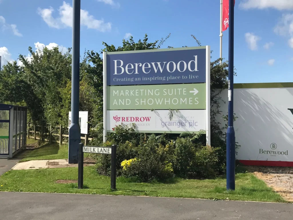 Sign for Berewood housing development with marketing suite and show homes directions. Surrounded by greenery, with a blue sky above