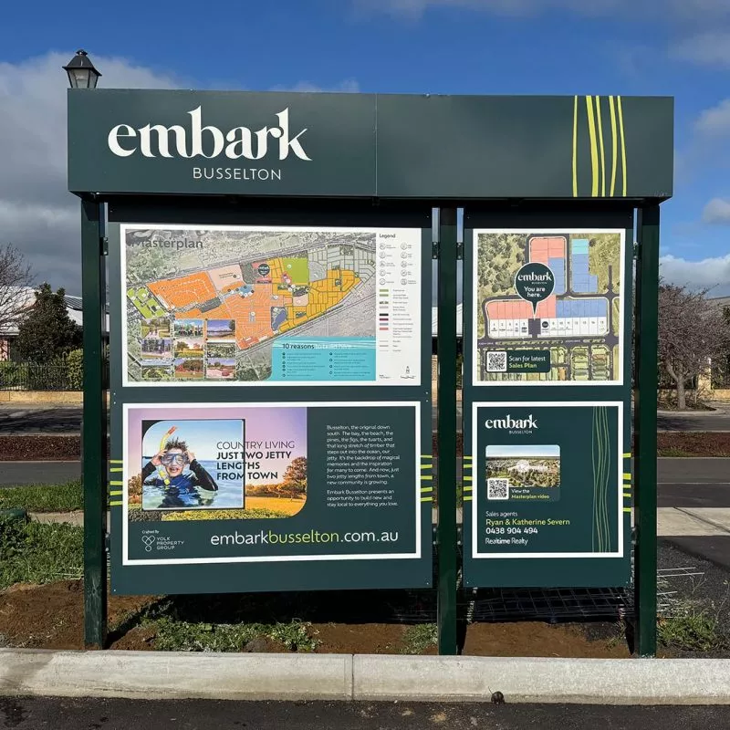 Outdoor sign for Embark Busselton featuring colorful site maps and promotional text. Set against a clear blue sky with surrounding greenery