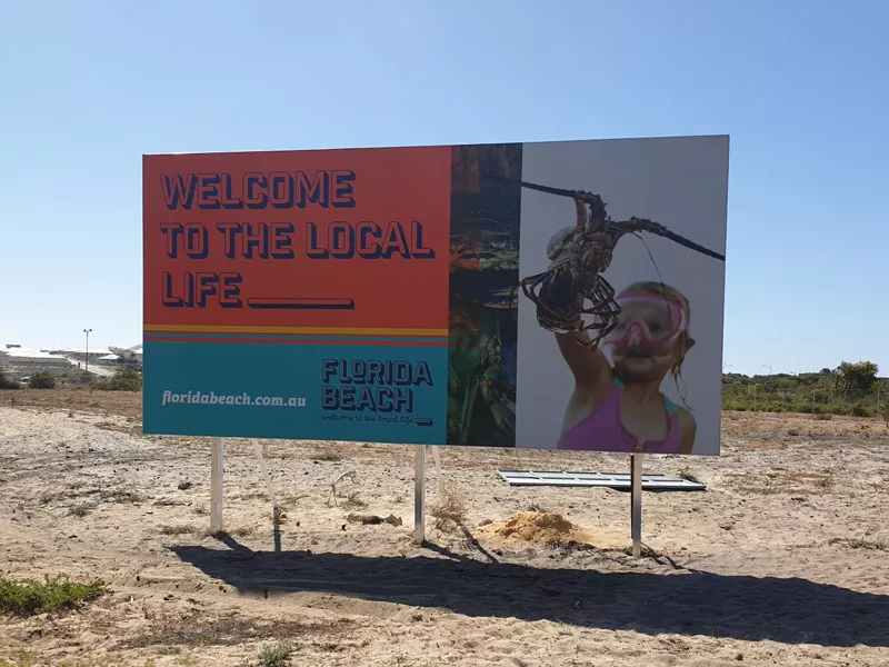 Billboard in sunny, sandy area reads “Welcome to the Local Life.” Image shows a child holding a large crab. Text: FLORIDA BEACH flordabeach.com.au.