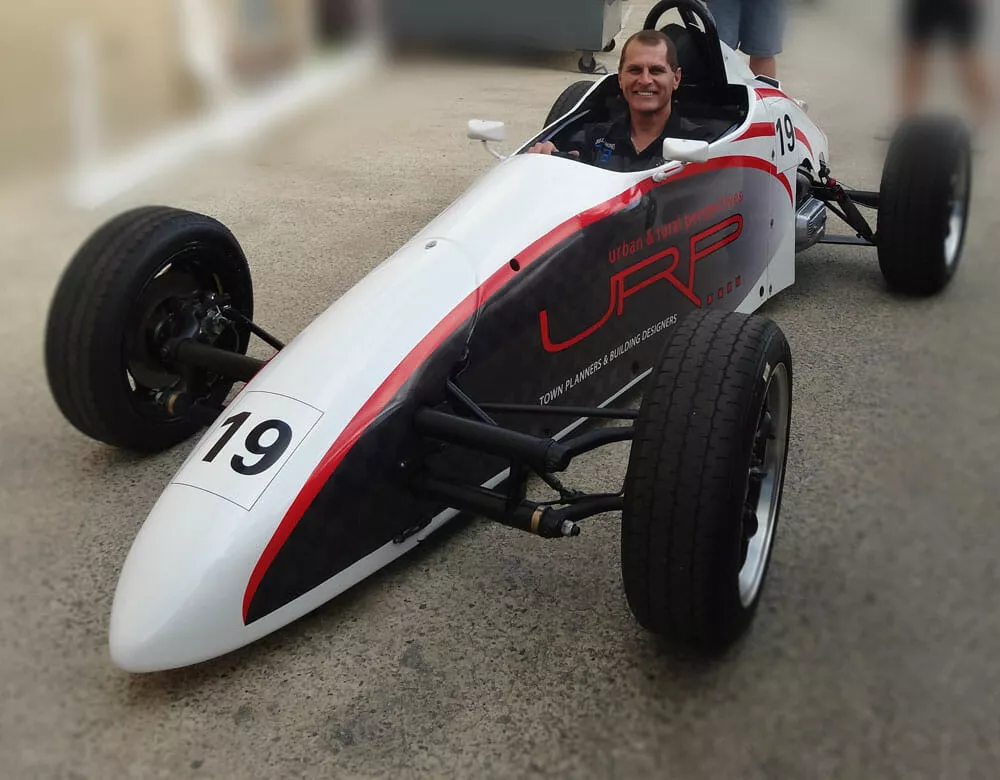 Smiling person sitting in a sleek white and black single-seater race car with number 19, parked in a pit lane, conveying excitement and power