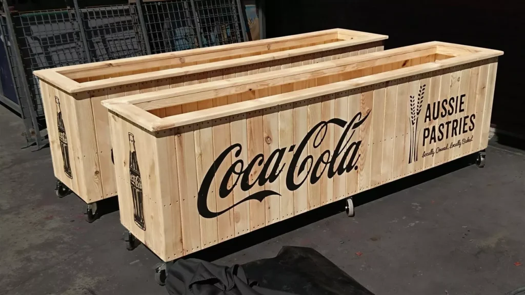 Wooden planters with logos for Coca-Cola and Aussie Pastries on the side. The planters are on wheels, set on a concrete floor, suggesting a rustic or industrial theme