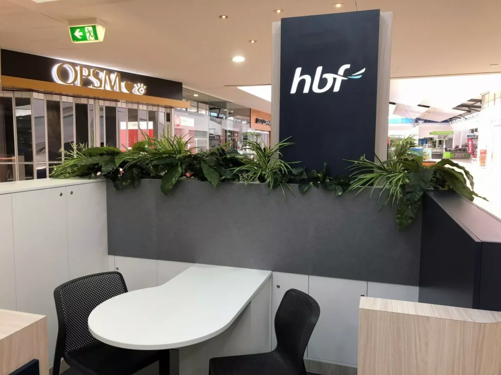A small seating area in a shopping mall, featuring a white table with black chairs. The surrounding divider displays the 'hbf' logo above decorative green plants. Nearby, the OPSM store is visible, .
