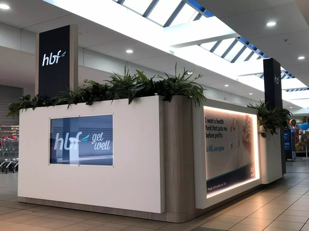 Modern kiosk in a shopping mall with "hbf" branding on signs and digital displays. Features decorative plants on top.