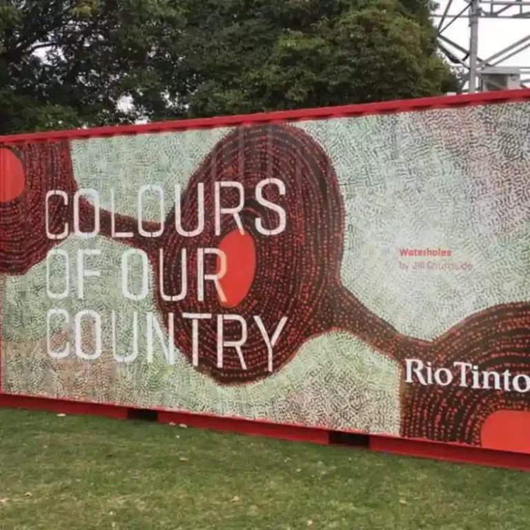 Outdoor art display featuring "Colours of Our Country" in bold text on a red, black, and white dotted pattern, with trees and grass in the background.