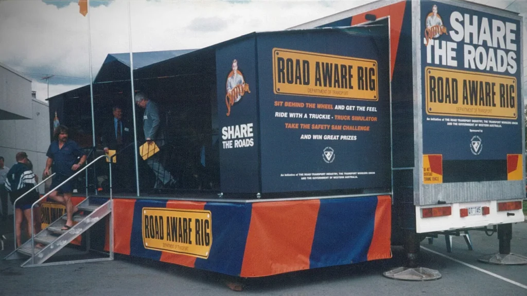 A promotional truck display for road safety awareness, featuring bold yellow "Road Aware Rig" signs. People are entering via steps. The tone is educational and engaging