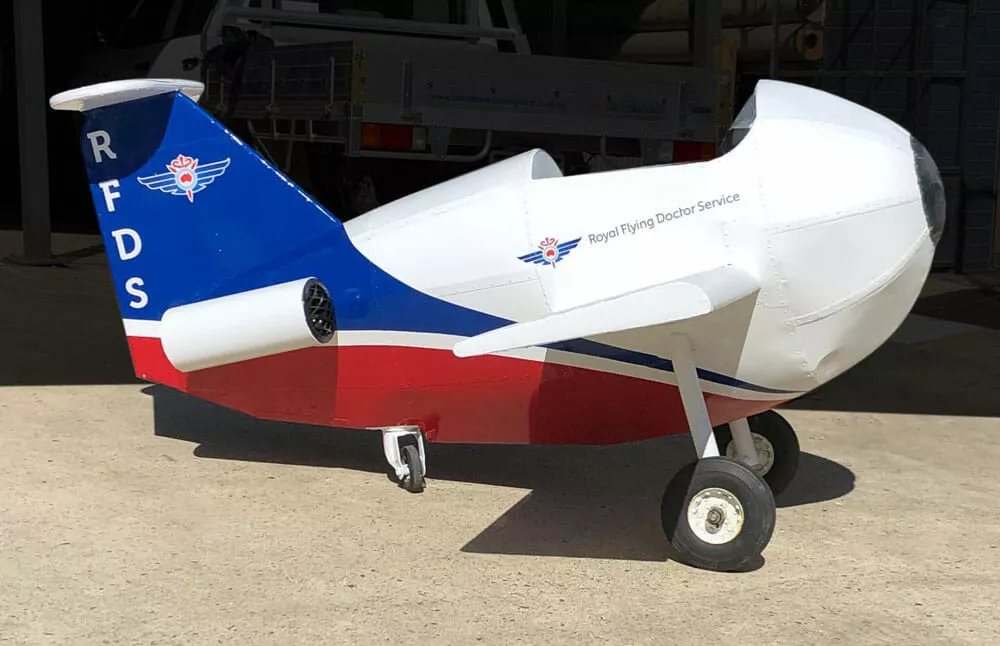 A small white and blue toy plane with "Royal Flying Doctor Service" logo on the side, sunlight casting a shadow on the ground, parked on concrete