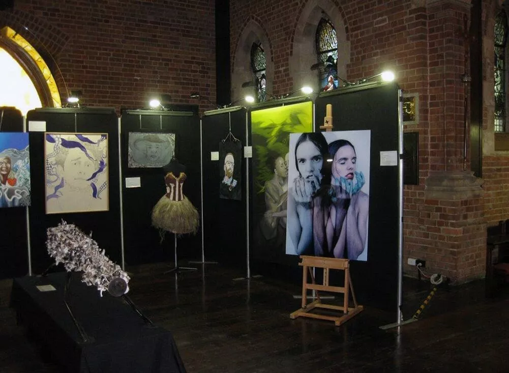 Art exhibition in a dimly lit room with brick walls. Black panels display various artworks, including paintings and a tutu. The atmosphere is creative and contemplative