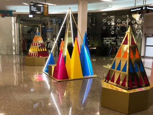Colorful geometric sculptures with triangles in yellow, blue, pink, and orange on gold bases are displayed in a glossy mall atrium
