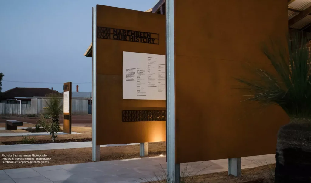 A modern memorial with rust-colored panels and text "We Remember Our History" under a dim evening sky