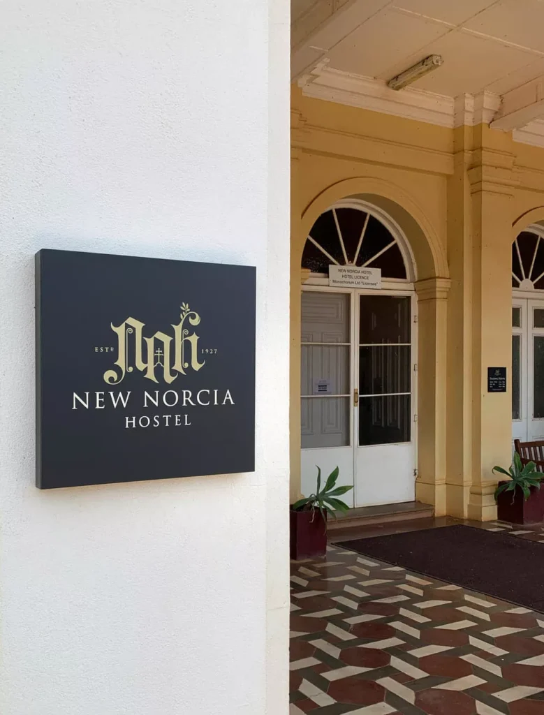 Sign for New Norcia Hostel with ornate lettering is mounted on a cream building. Arched doorways and potted plants create a welcoming entrance