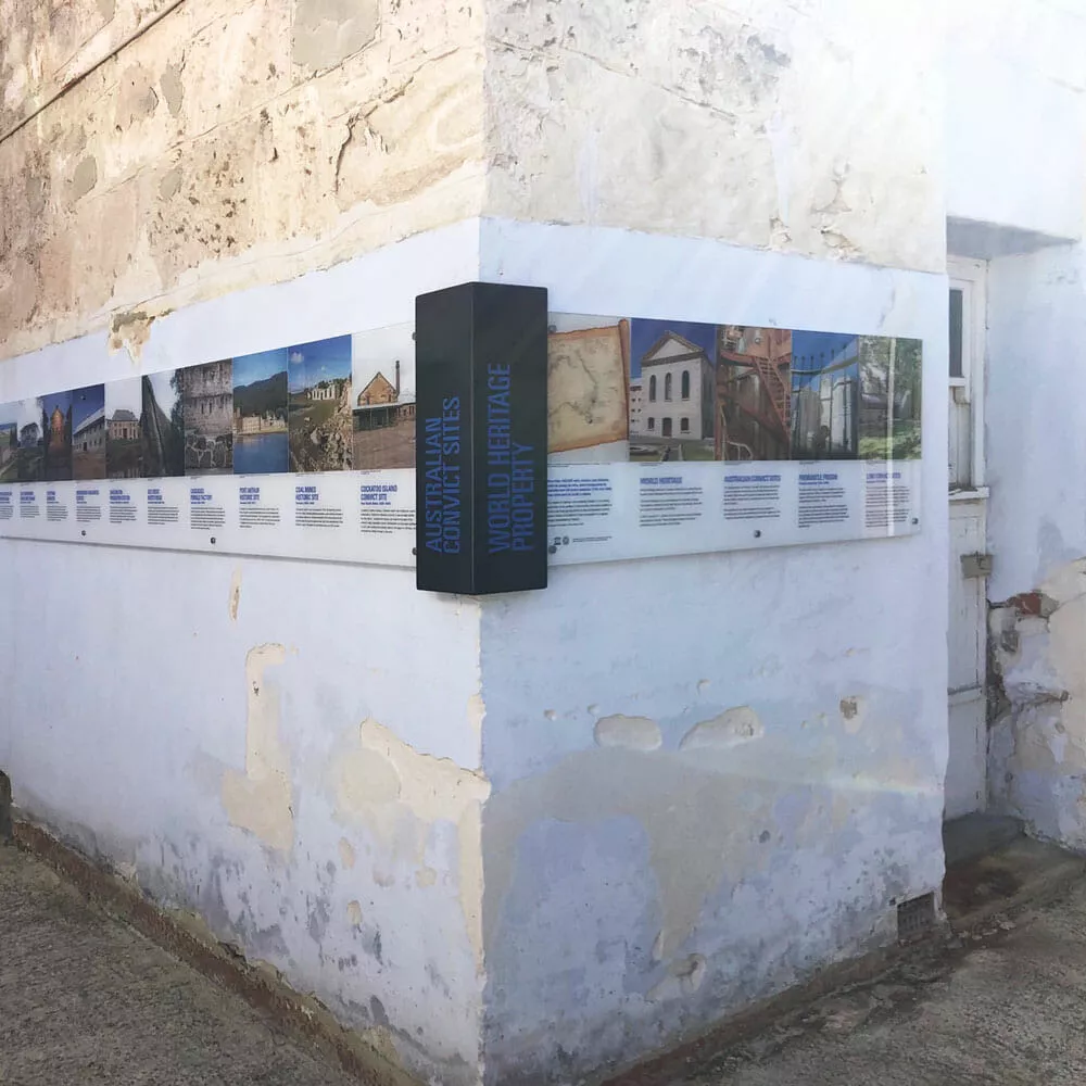 A weathered stone wall corner displays a sign about "Australian Convict Sites," featuring images and descriptions. The scene conveys historical significance