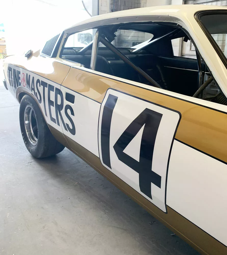 A vintage race car with the number 14 and the words "Line Masters" on the side. The car is gold and white wrapped, parked in an indoor garage