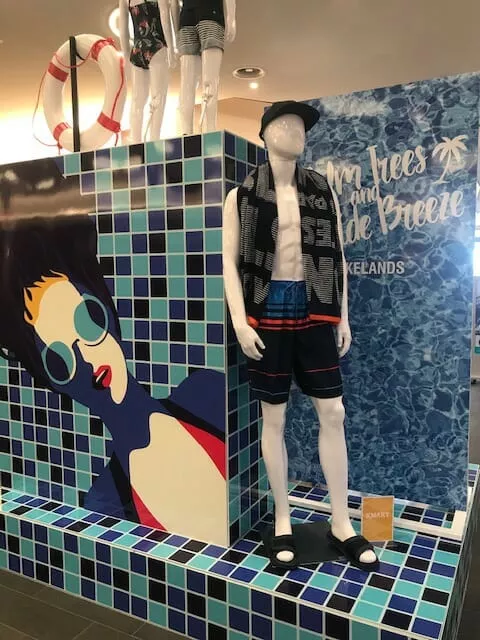 Mannequin in swim trunks, sandals, and a hat stands near a blue-tiled wall with beach-themed graphics. Energetic, summery display ambiance