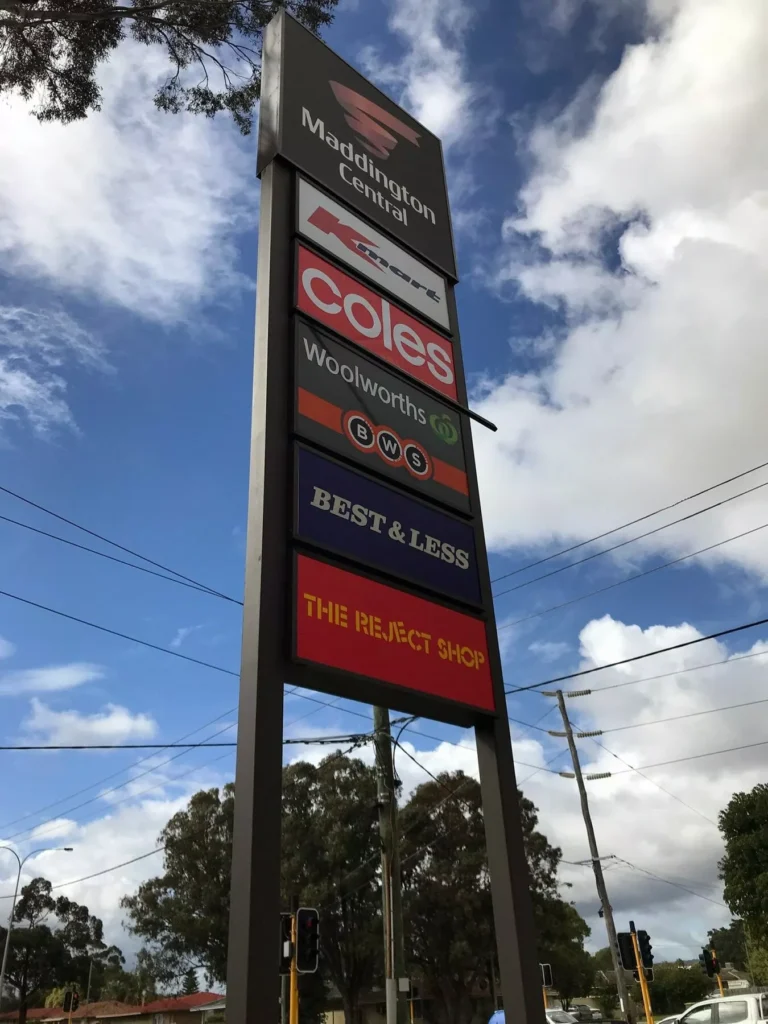 Signboard of Maddington Central shopping center against a partly cloudy sky, displaying logos of stores like Coles, Woolworths, and The Reject Shop