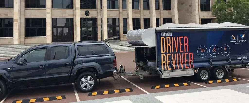 A dark blue pickup truck towing a trailer marked "Driver Reviver" is parked in front of a large building with stone columns, conveying a supportive community service