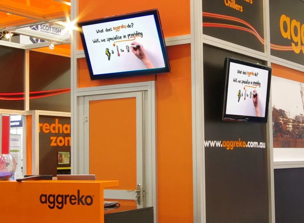 Trade show booth for Aggreko featuring orange branding. Two screens display cartoon hands writing text