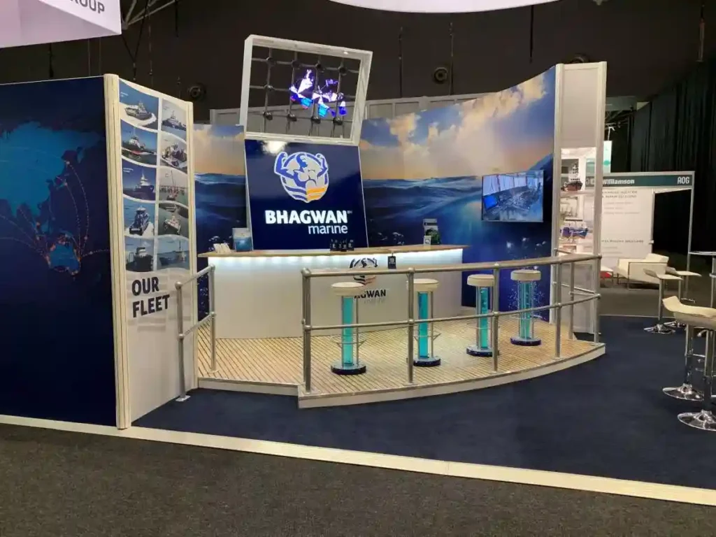 Exhibition booth with “Bhagwan Marine” logo, nautical backdrop, and three stools. Displays highlight marine fleet; the atmosphere is professional and inviting.