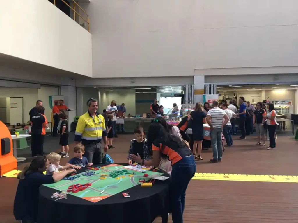 A lively indoor event with adults and children engaging in tabletop games and activities. People are casually dressed, creating a relaxed, friendly atmosphere.