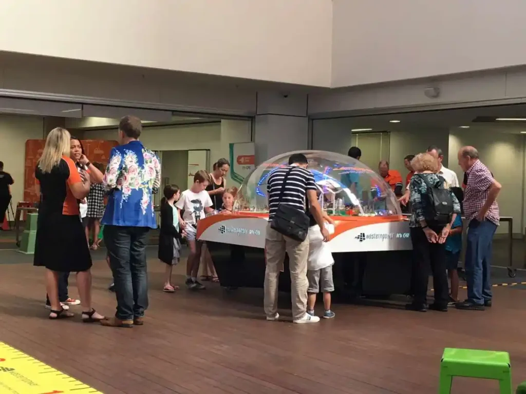 People of various ages gather around a large, colorful interactive science exhibit under a clear dome. The scene is lively and engaging.