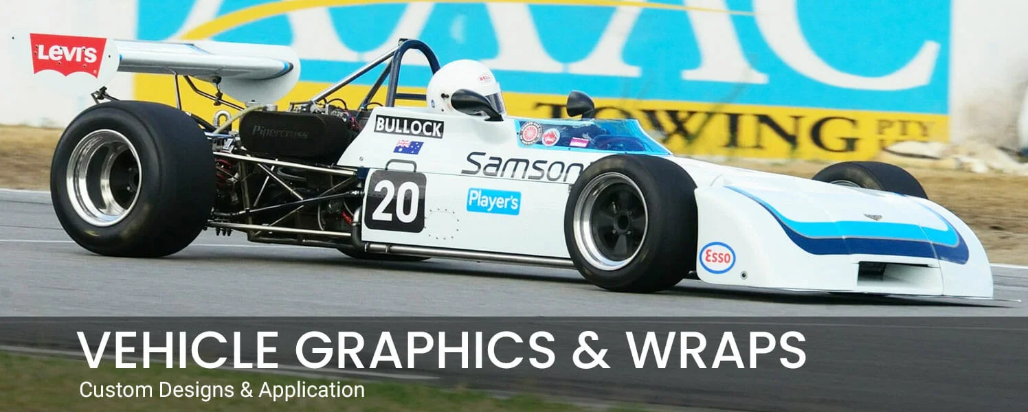A white classic open-wheel racing car on a track, featuring custom vehicle graphics and sponsor decals. Overlay text reads: 'Vehicle Graphics & Wraps - Custom Designs & Application'.