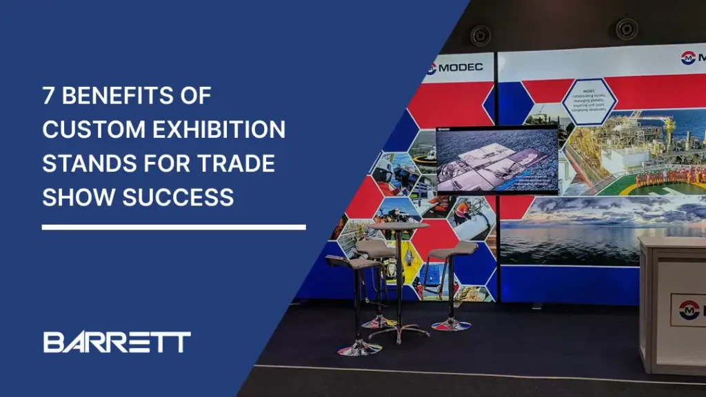 Trade show booth with bold red and blue geometric design featuring industrial images. Text overlay reads '7 Benefits of Custom Exhibition Stands for Trade Show Success.' Two modern stools and a high table are in front. The tone is professional and sleek."